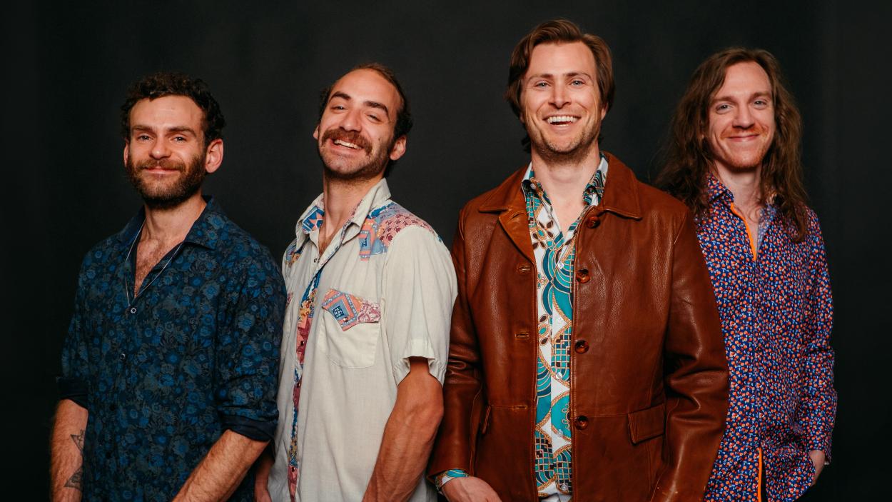Four smiling light skinned men stand against a black backdrop.