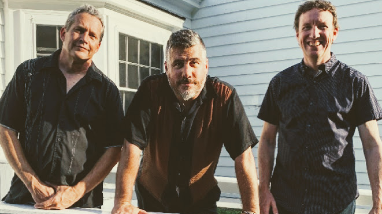 Three light skinned men stand together in front of a house with white siding