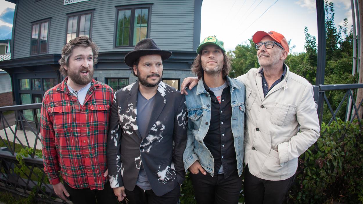 Four light skinned men stand with their arms around each other with a grey house and trees behind them.