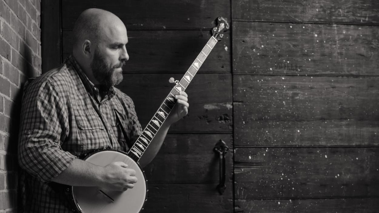 A black and white image of a light skinned man holding a banjo and leaning against a barn board wall