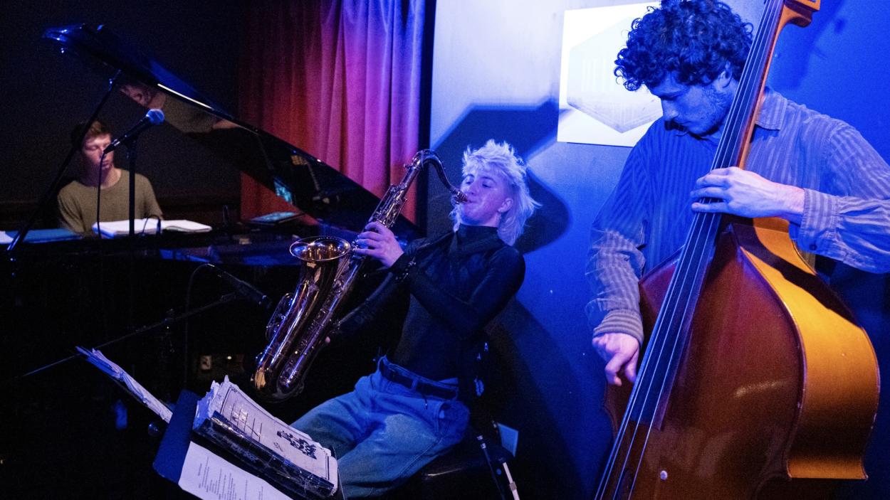 A light skinned man plays an acoustic double bass next to a light skinned woman playing a saxophone with a light skinned man playing a grand piano in the background. They are bathed in purple light