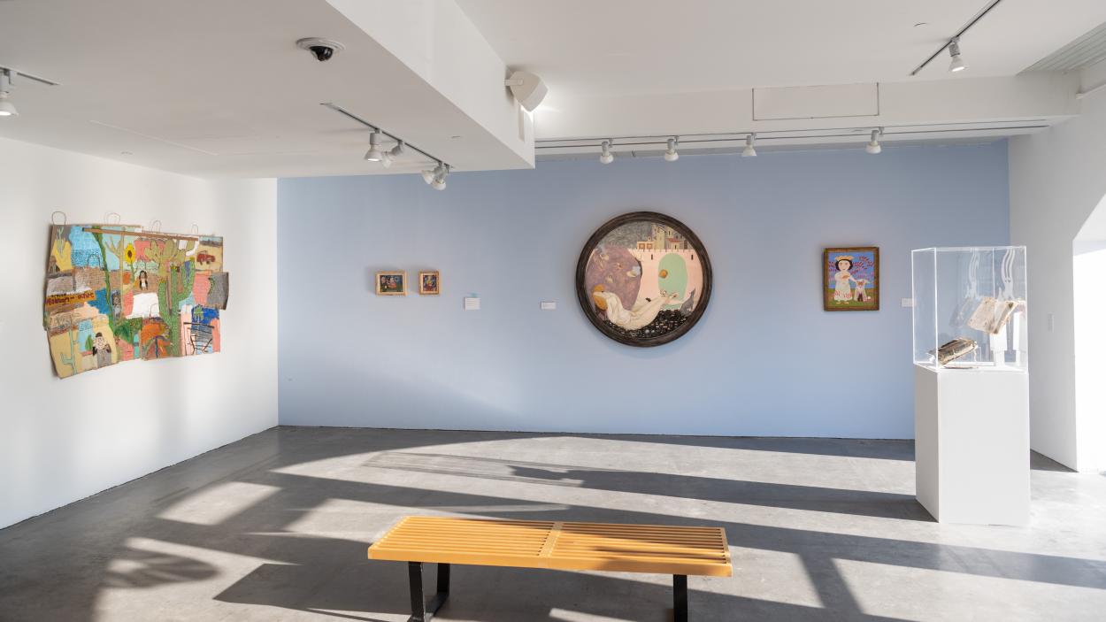 Paintings hang on the wall of an art gallery with one white wall and one light blue one. Light streams in from windows out of the frame and a blond wood bench and a sculpture under a vitrine on a white pedestal sit in the center of the room