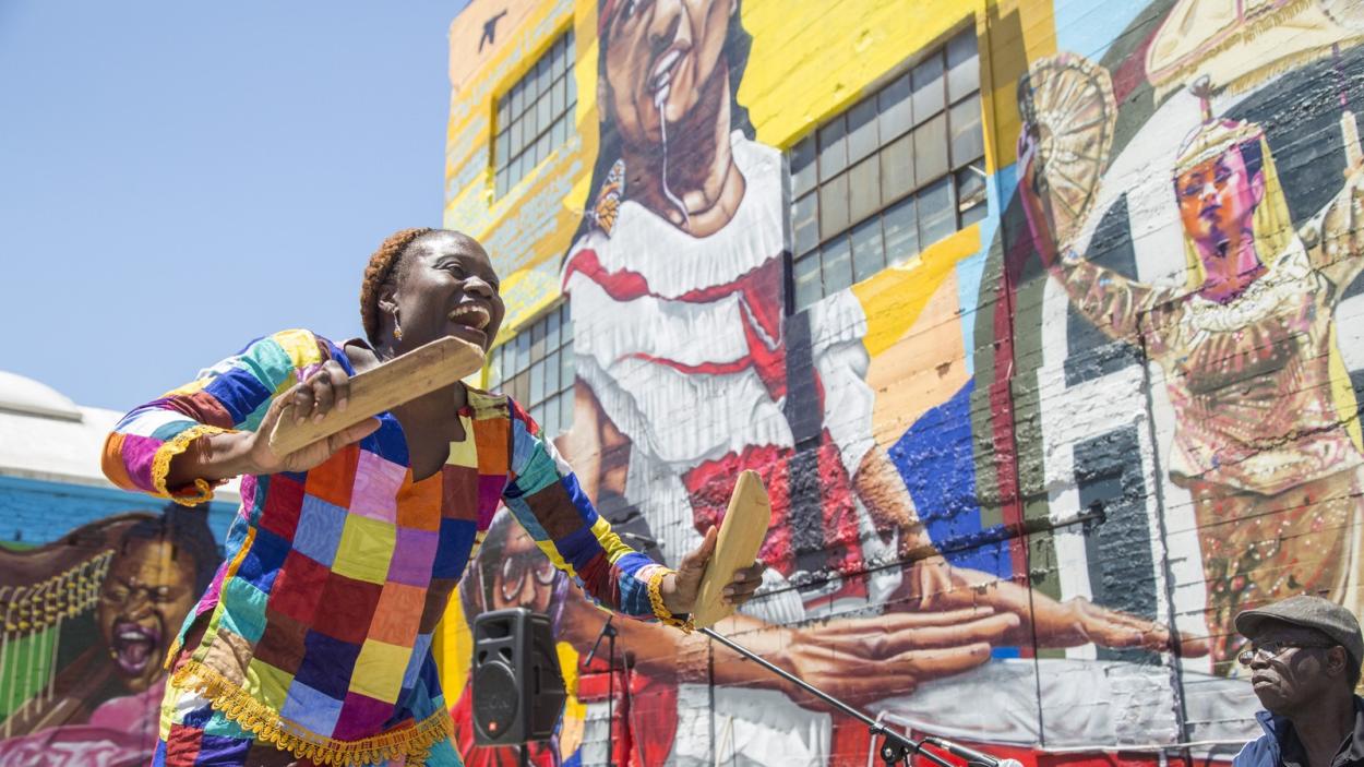 A photograph of a dark skinned woman wearing a multi colored blouse, holding two wide sticks as she smiles and dances in front of a large colorful, mural on a brick building