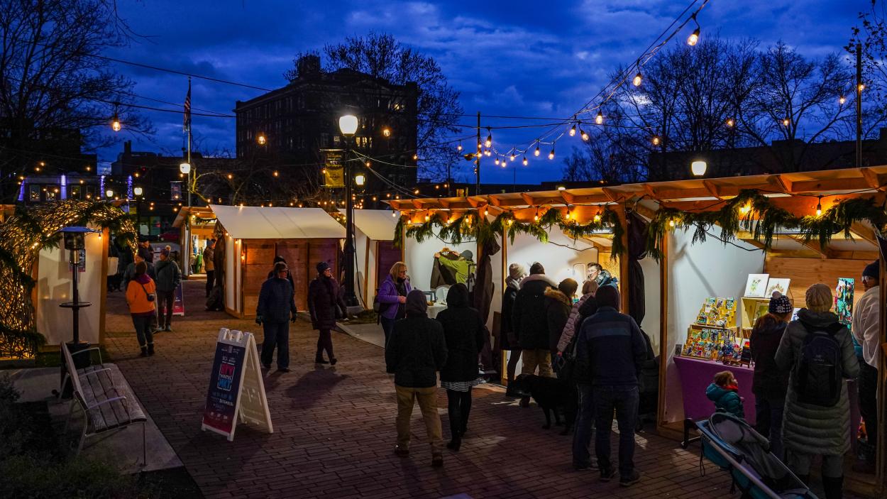 A photo of the BTV Winter Market at twilight. Wooden sheds decked with evergreen garlands and white twinkle lights are arranged around a brick lane. People wearing winter coats mill around and cluster round each vendor's booth, which are illuminated