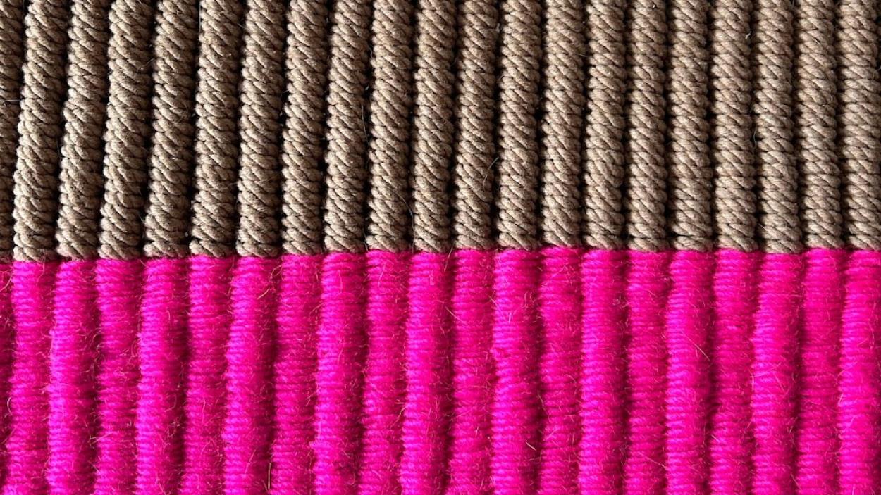 a photograph of a close up of a piece of textile art comprised of tightly woven ribs of thread. The top half are light brown and appear to be made of jute and the bottom half are bright magenta.