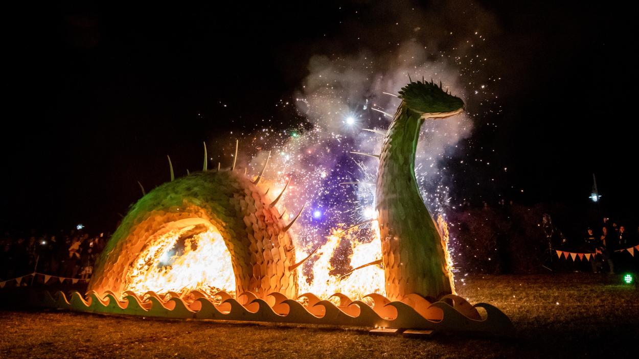 A photograph of a wooden sculpture a sea monster that has been lit on fire at night. Purple sparklers and fireworks shoot up from the flames and a crowd of people behind ropes strung with colorful flags is visible in the background