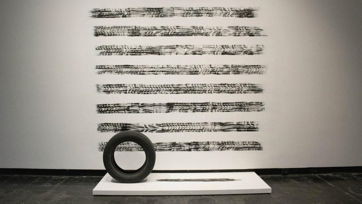 A photograph of a sculptural installation featuring a black tire on a low white platform, with 8 horizontal stripes of black tires tracks on the wall behind.