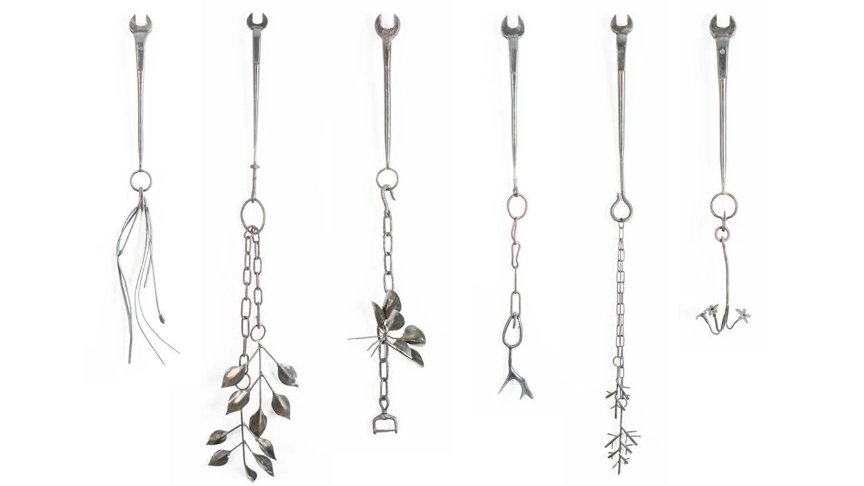 A photograph of six silver steel sculptures arranged vertically. Each work starts with a wrench that ends in a ring with danging chains that turn into flowers, vines, and other naturalistic abstract shapes
