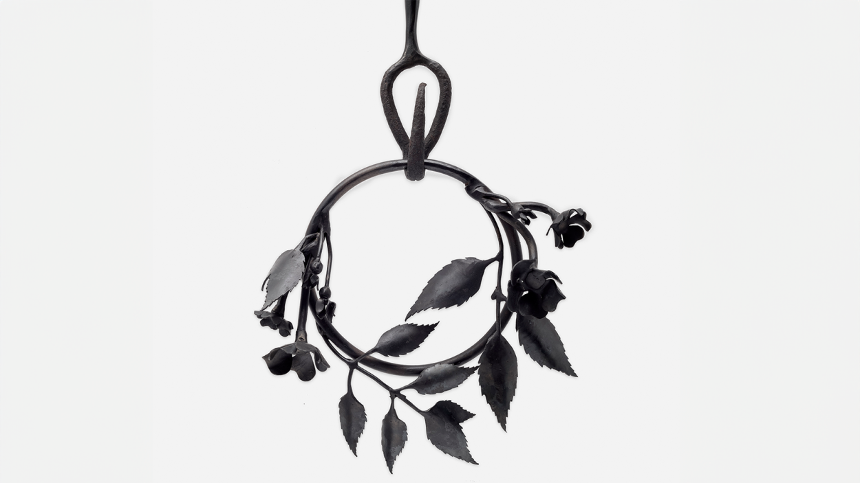 A photograph of a detail of a black steel sculpture that consists of a ring with leaves, flowers and vines twined around hanging from a hook a ring
