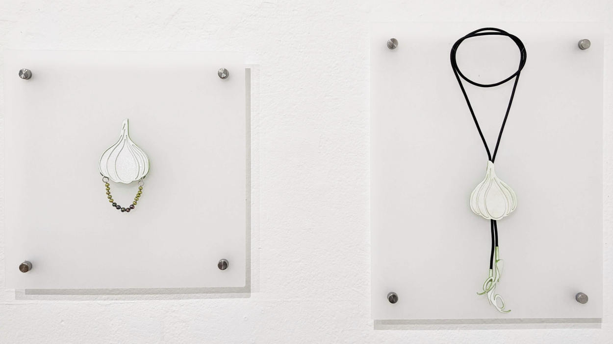 A photograph of two pieces of jewelry, mounted on a white wall. On the right is a brooch with a white bulb of garlic with a delicate chain looped to the bottom side. On thee right is a bulb of garlic hanging from a brown leather string looped around several times