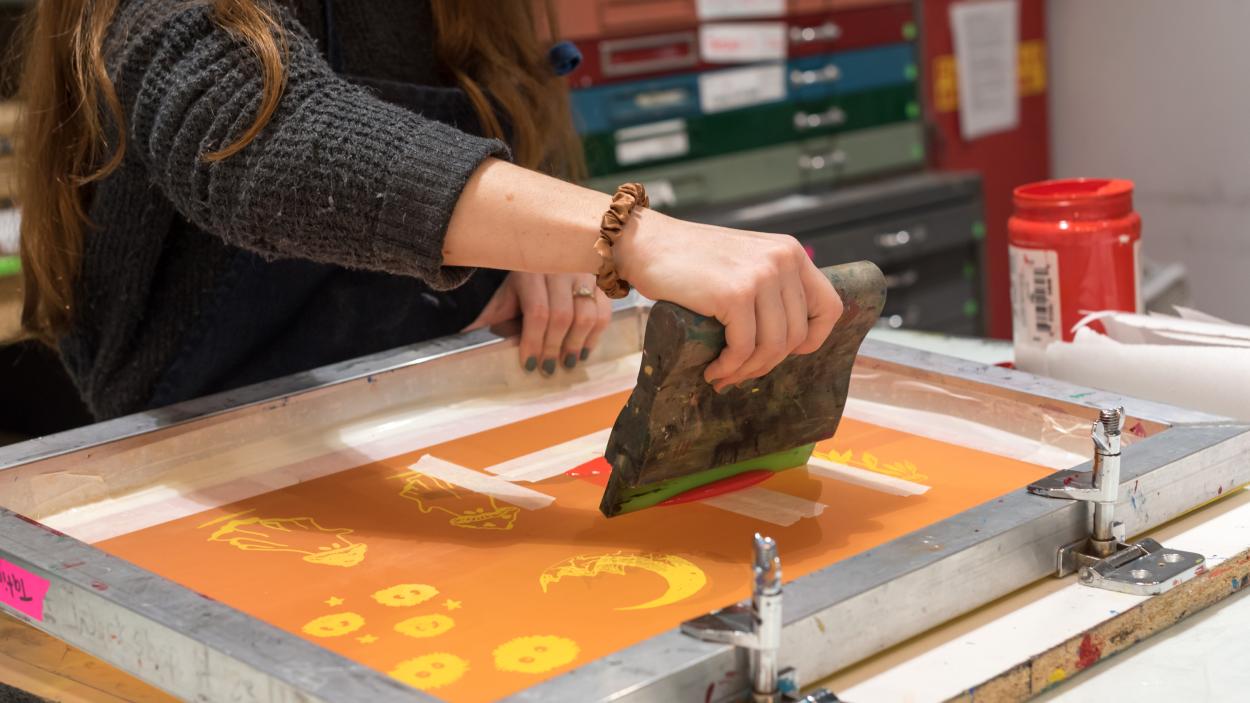 A close up photograph of a light skinned woman's hands putting ink on a screen for screen printing. The screen has an orange background with yellow designs burned out