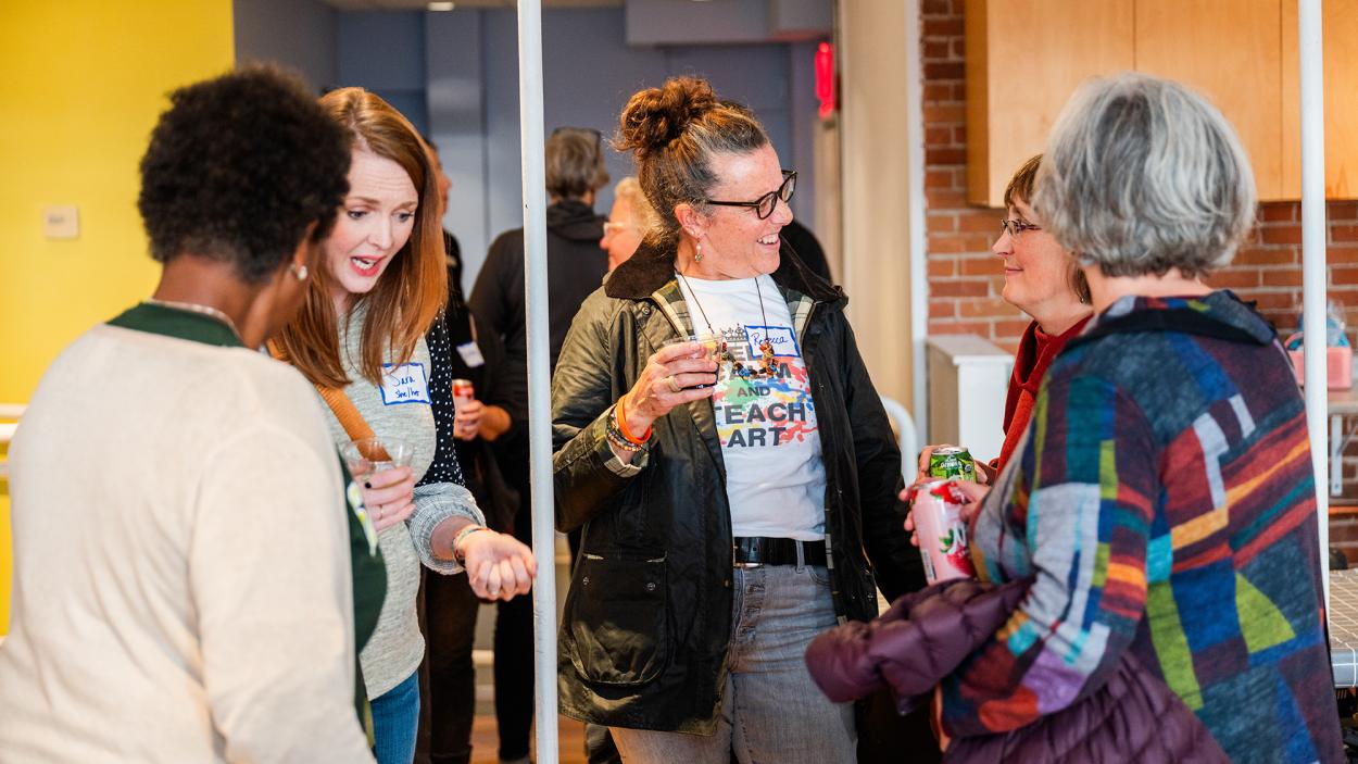 A photo of five women standing together and talking in the BCA Center Gallery Learning Studio Space. One woman has dark skin and short hair and stands with her back to the camera. One has light skin and reddish brown straight hair and exclaims while holding a plastic cup. In the center of the group a woman with greyish brwn hair in a bun and light skin wears a t-shirt that says "Keep Calm and Teach Art" while holding a plastic cup and laughing