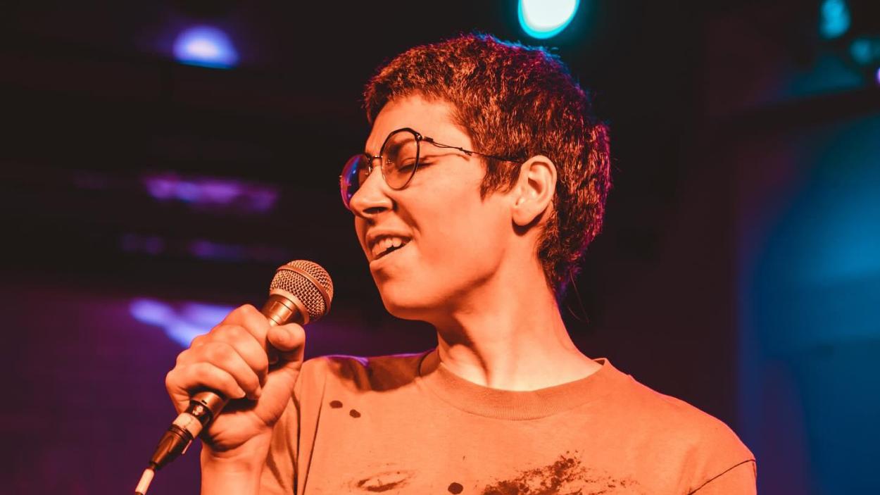 A person with short hair and glasses singing into a microphone.