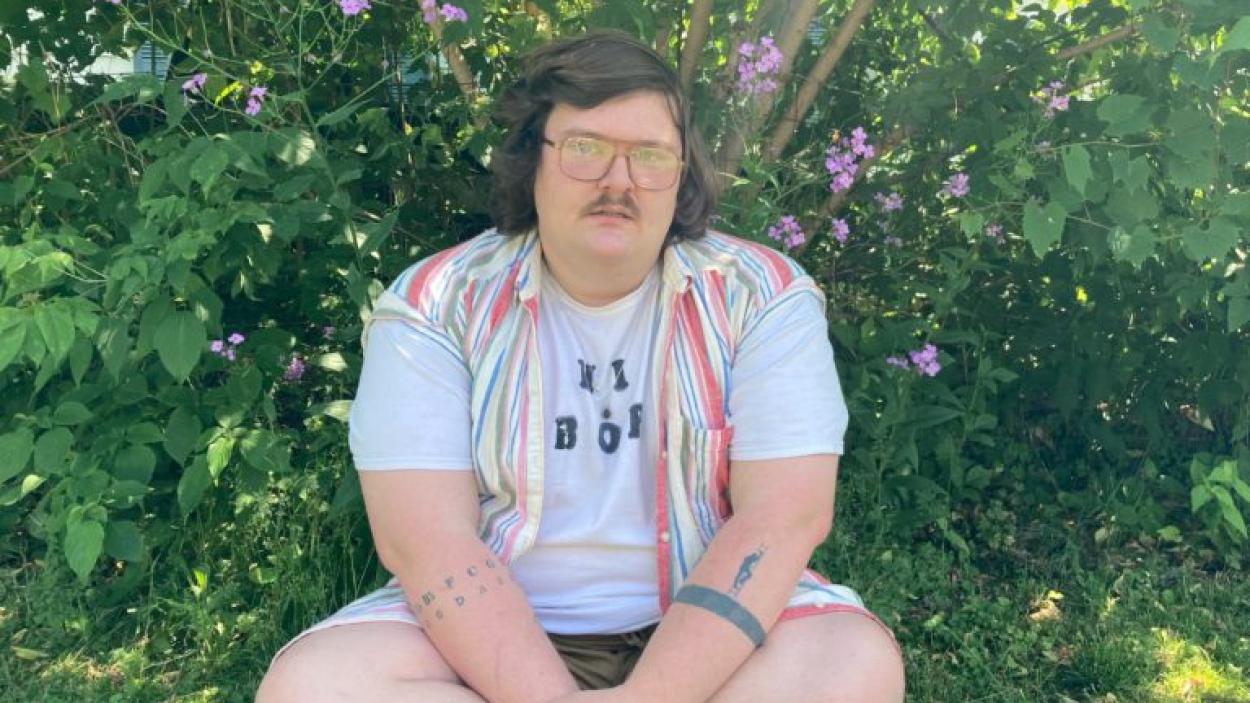 A person with glasses and a mustache and wearing a vest sits against a backdrop of green grass and bushes