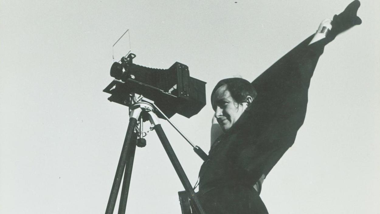 A black and white image of a woman standing behind a camera on a tripod.