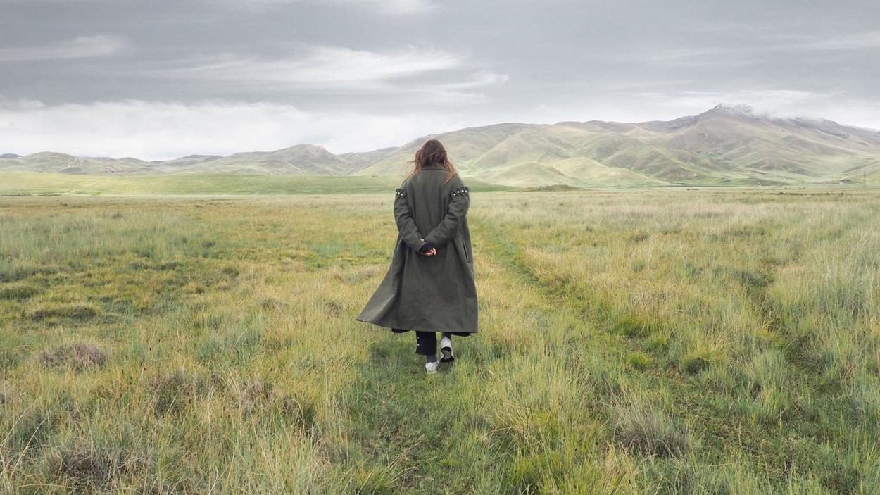 An image of a person in a long coat with their back to the camera, stranding in a mountainous meadow.