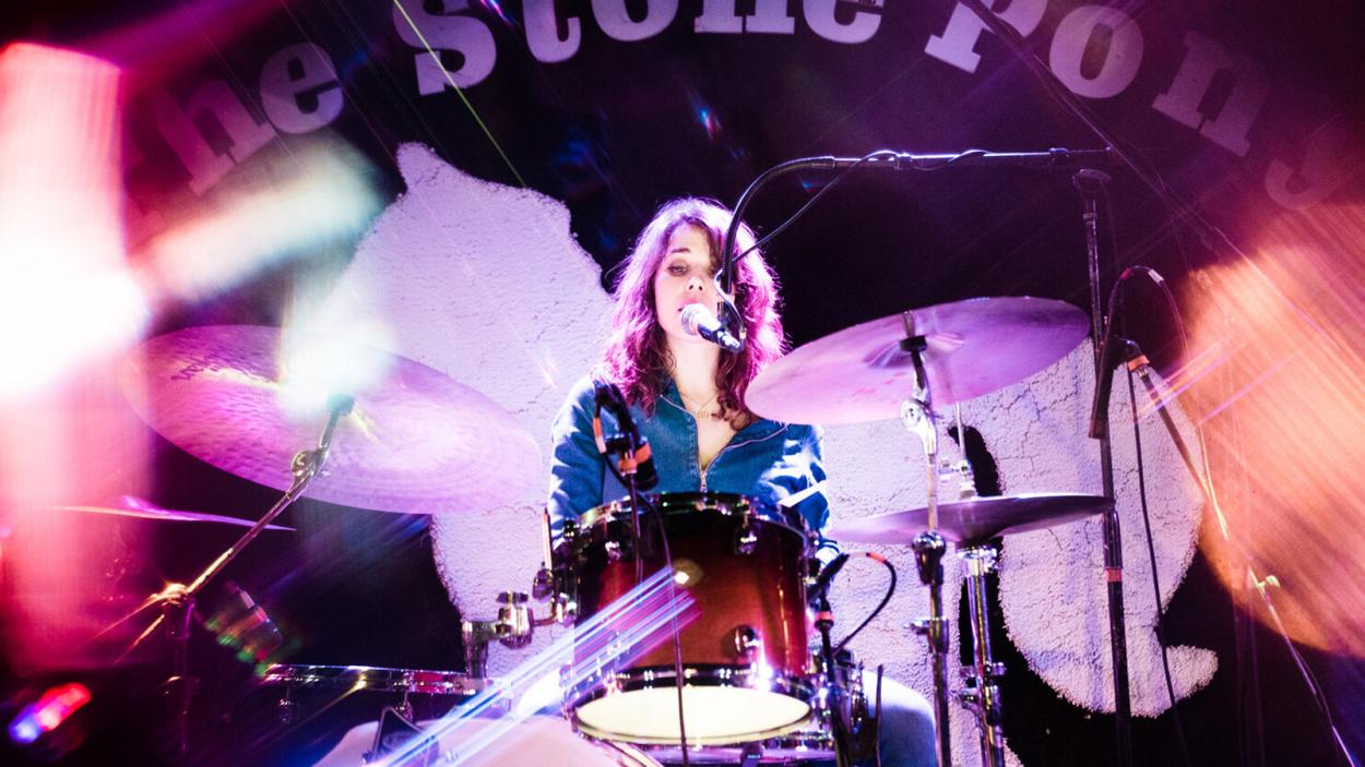 the performer sitting behind a drumkit singing into a microphone. Photo has some beautiful layered lighting effects