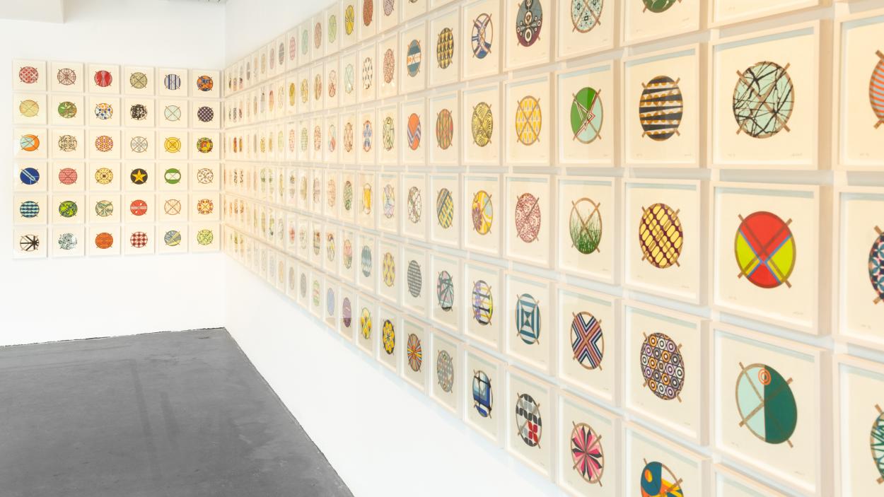 a photograph of an installation in the BCA Center gallery by Jacob Hashimoto made up of hundreds of framed circular, colorful prints, hung in a grid that wraps around a corner of the gallery wall
